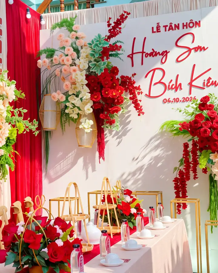 Phông lễ tân hôn với chữ hỷ đỏ, rèm trắng và các cụm hoa lụa đỏ, kem do Mỹ Liên trang trí